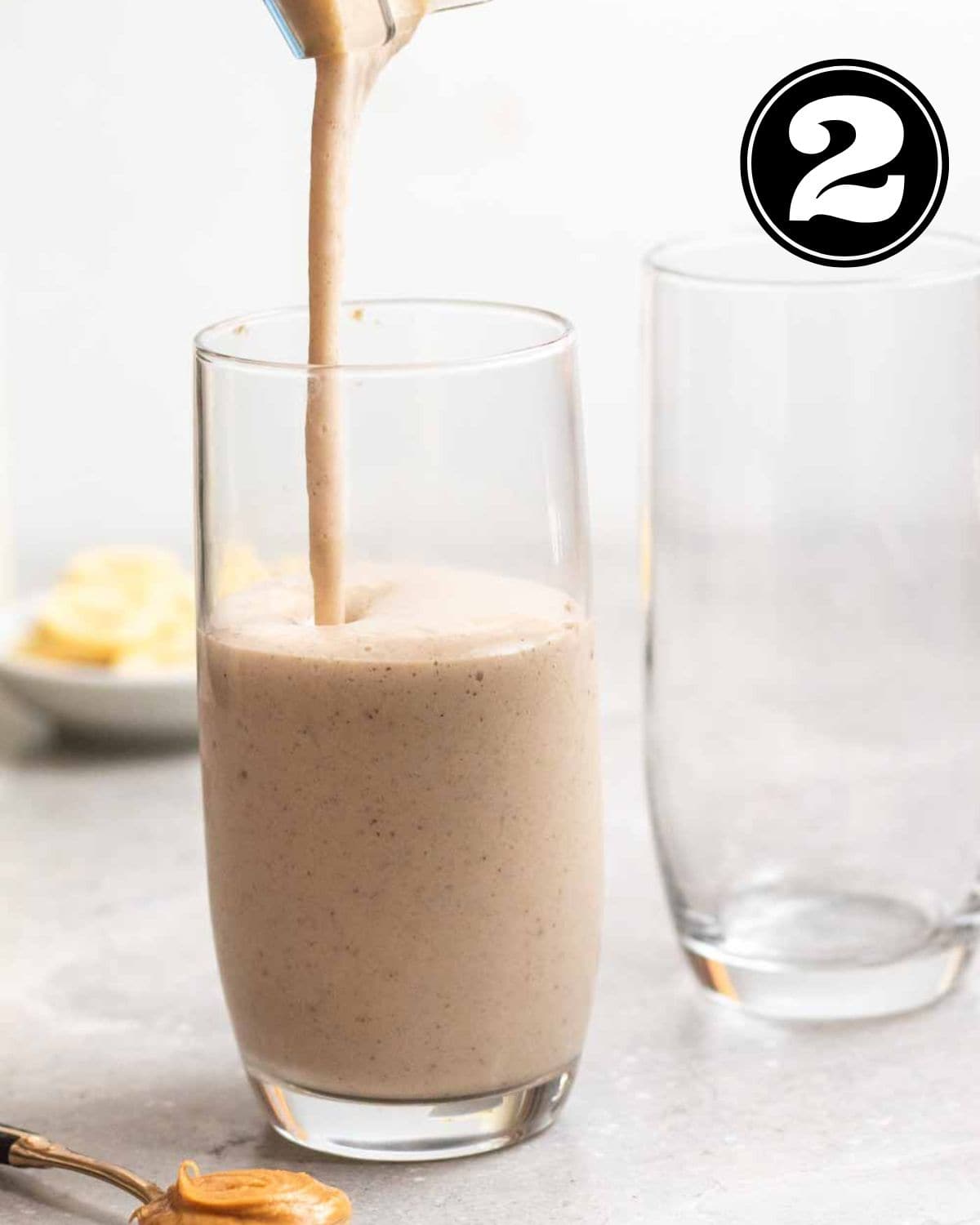 Pouring smoothie into a glass with another empty glass and banana slices in the background.