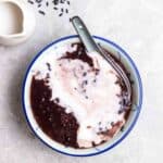 A bowl of black glutinous rice dessert served with spoon with coconut milk beside.