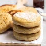 A stack of crumpets on a wooden board with more crumpets in the background.
