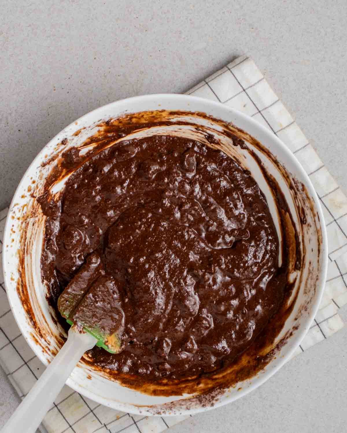 Brownie batter in a bowl with a spatula beside on a folded white cloth.