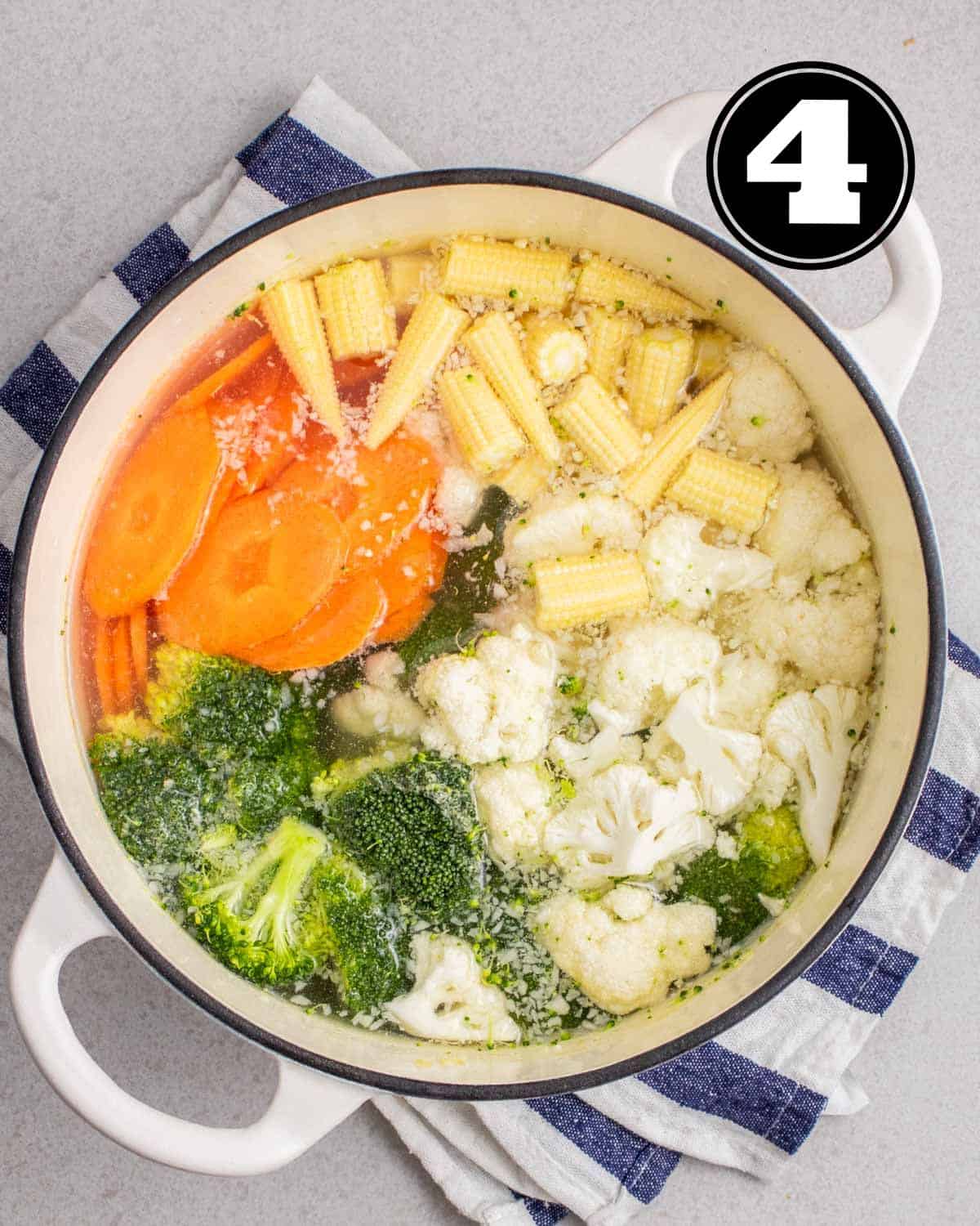 Uncooked vegetables in broth in a white Dutch oven on a white and blue folded teacloth.