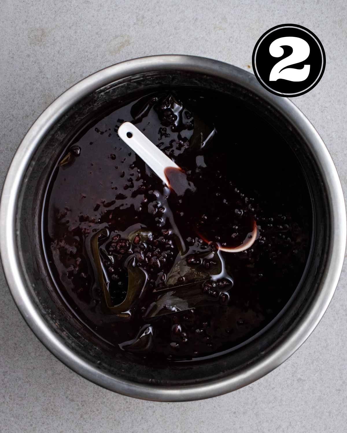 Cooked black glutinous rice dessert in a pot with a white ceramic spoon inside.