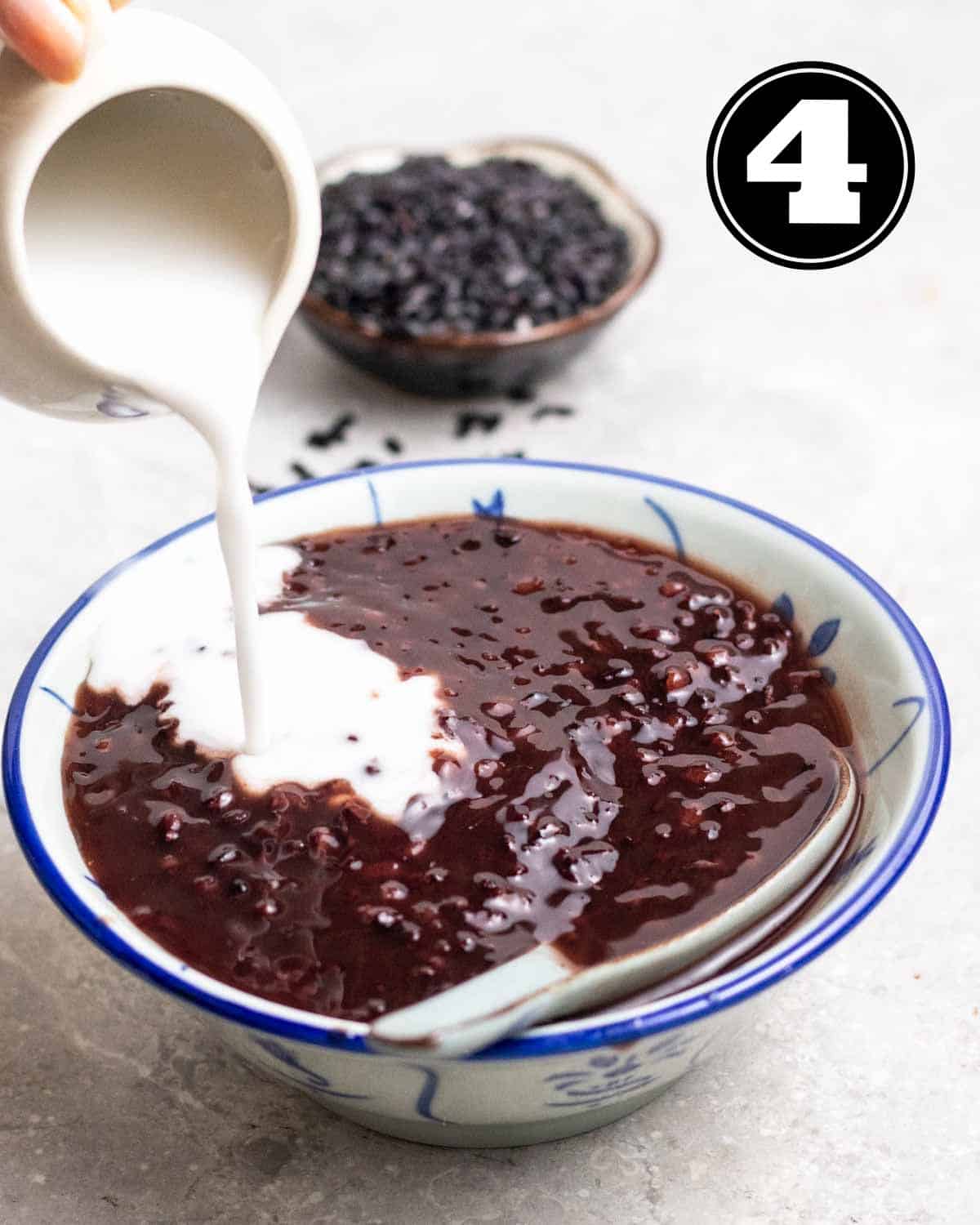Pouring coconut sauce over a bowl of black glutinous rice dessert served with ceramic spoon.