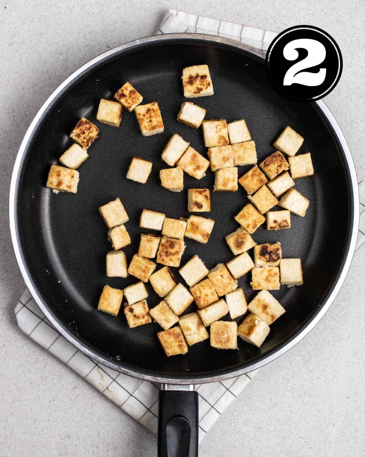 Pan-fried tofu in a non-stick pan on a folded white checkered teacloth.