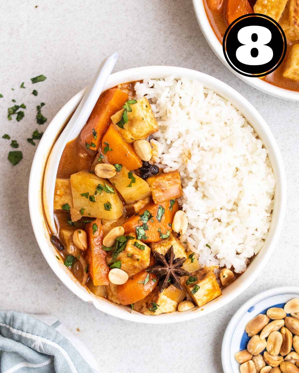 A bowl of Massaman curry and rice served with a ceramic spoon with another bowl of curry and peanuts beside.