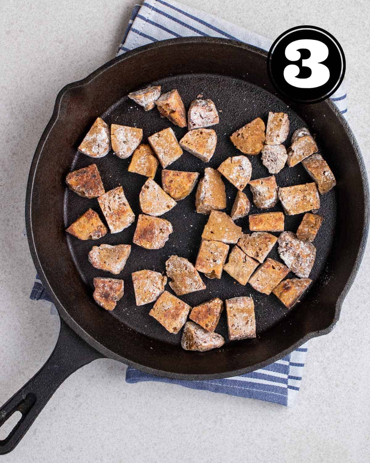 Fried seitan cubes in a cast iron skillet on a blue folded teacloth.