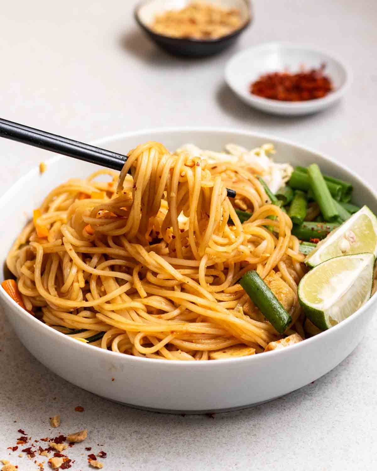 Taking out some noodles from a plate of pad Thai with chili flakes and chopped peanuts in the background.