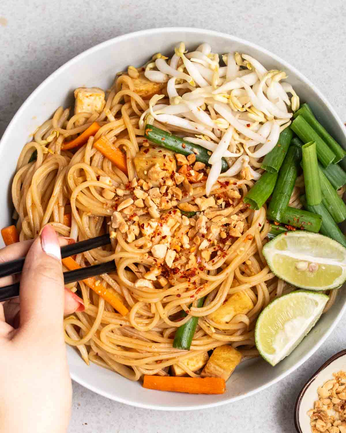 Stirring a plate of tofu pad Thai with chopsticks served with bean sprouts, lime wedges, and green onion.