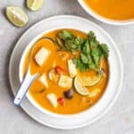 A bowl of tom kha on a plate served with a ceramic spoon with lime wedges and another bowl of curry beside.