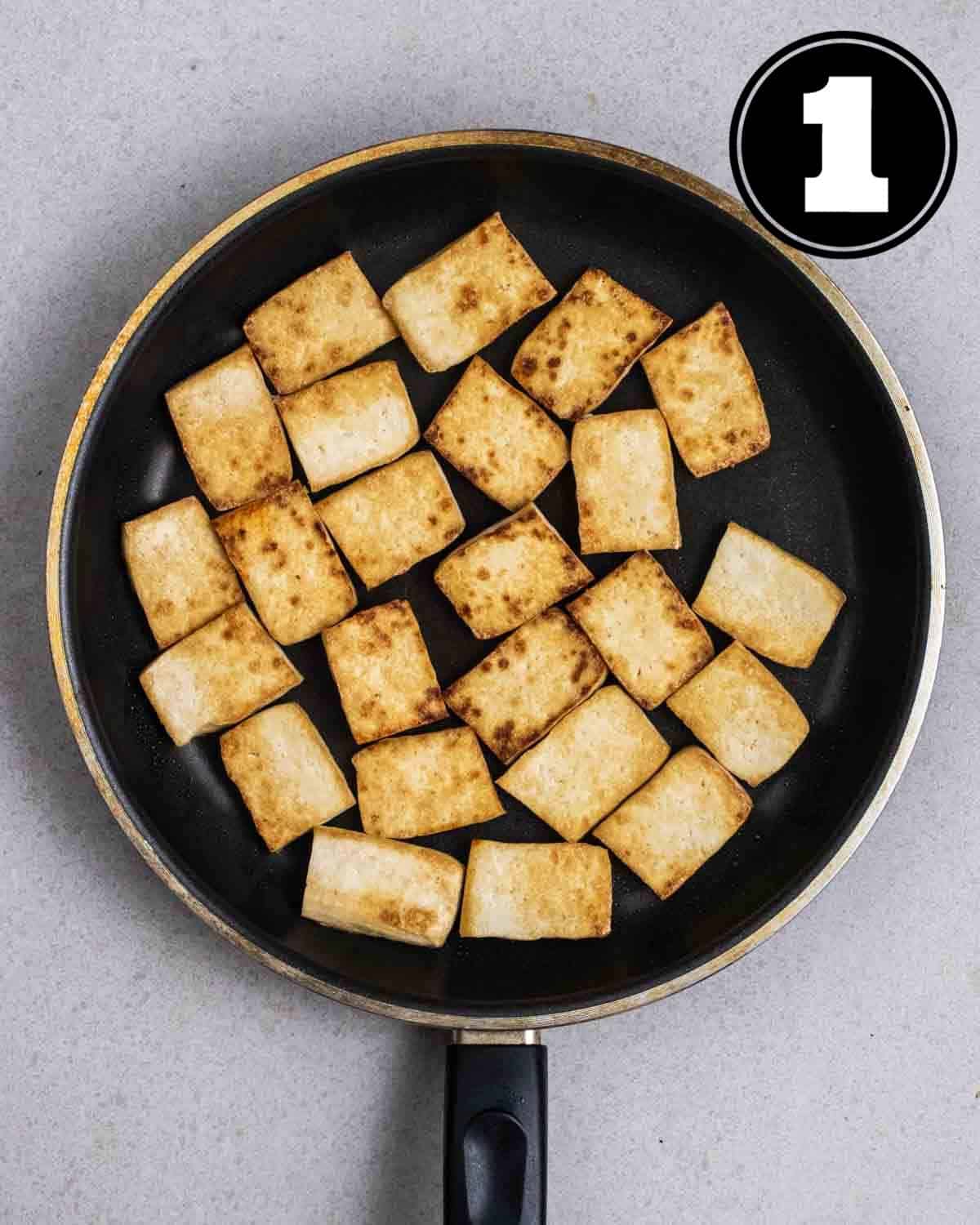 Pan-fried tofu in a non-stick pan on a grey background.