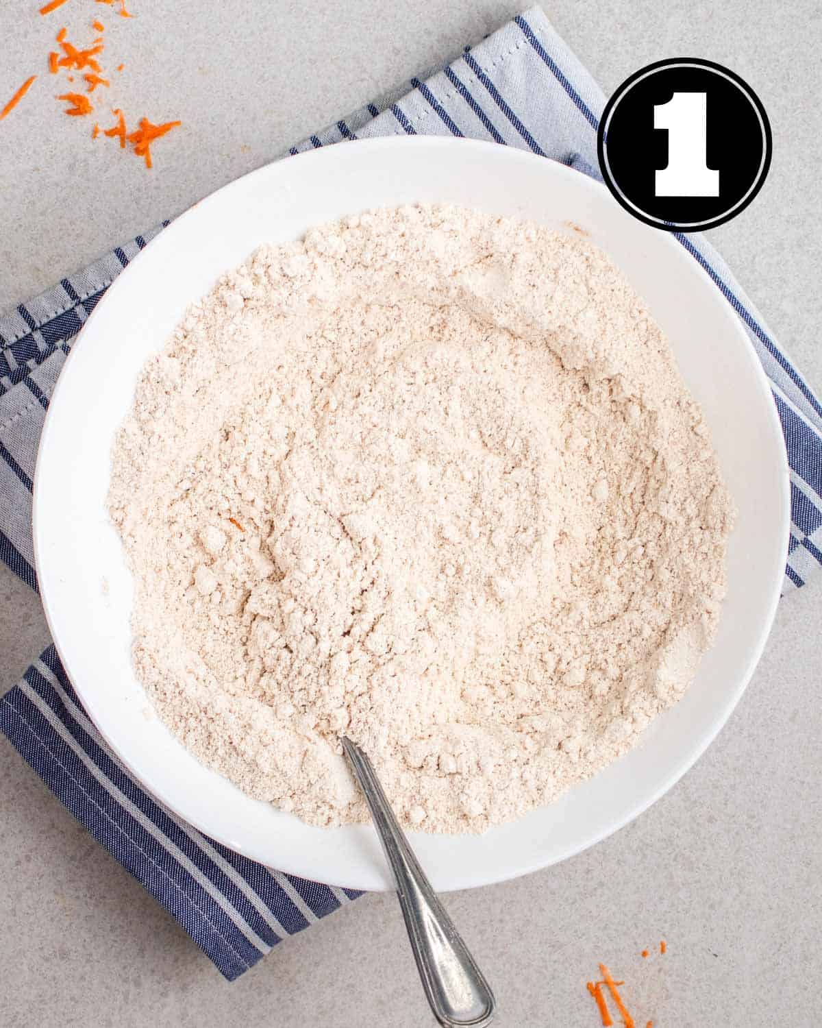 Dry ingredients for applesauce carrot muffins in a large white bowl with a spoon beside.