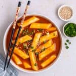 A bowl of tteokbokki served with chopsticks with chopped green onion and sesame seeds beside.