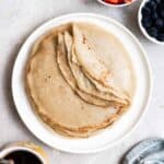 A stack of crepes on a plate with strawberries, blueberries, and coffee beside.