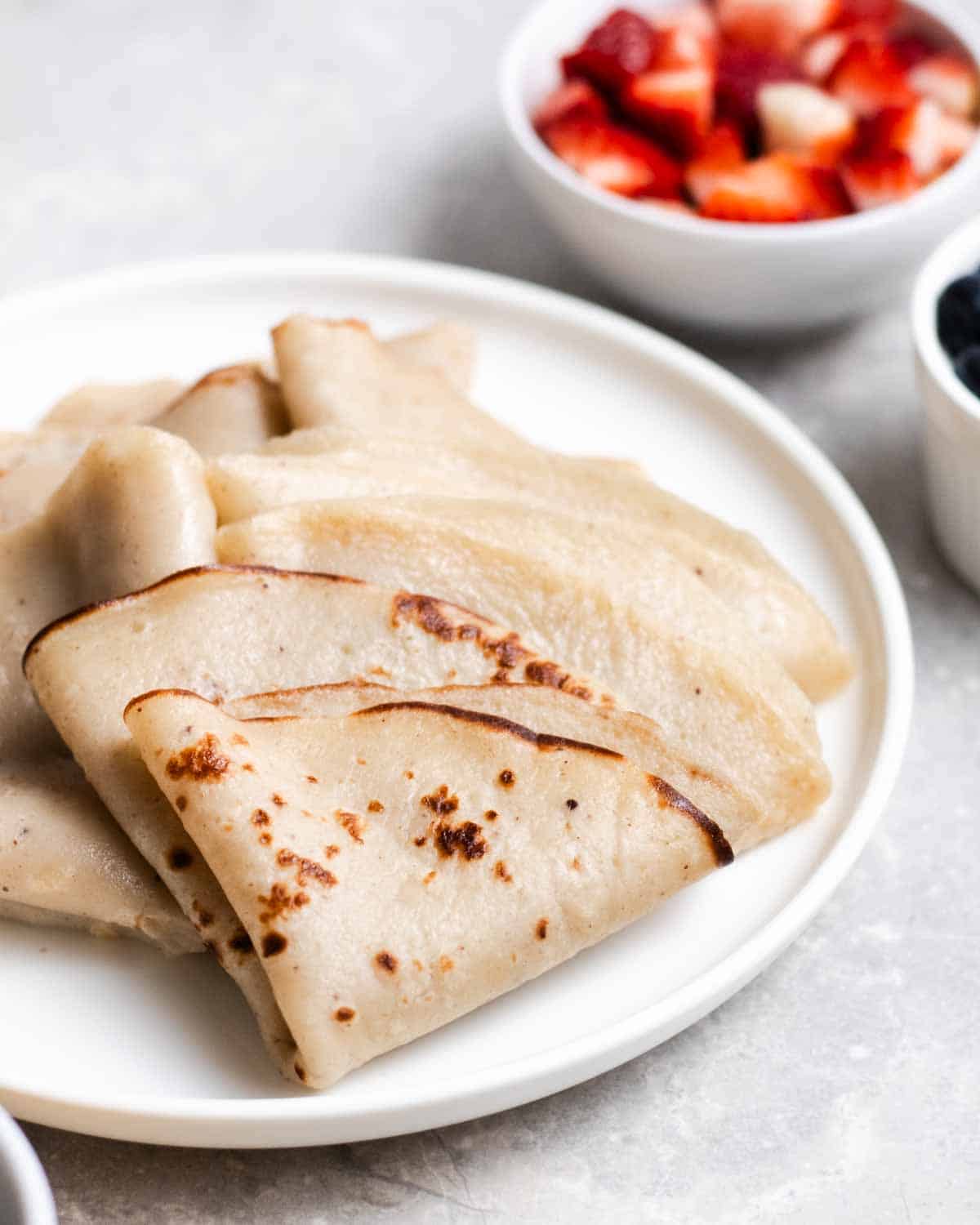 A plate of crepes folded into triangles with blueberries and chopped strawberries in the background.