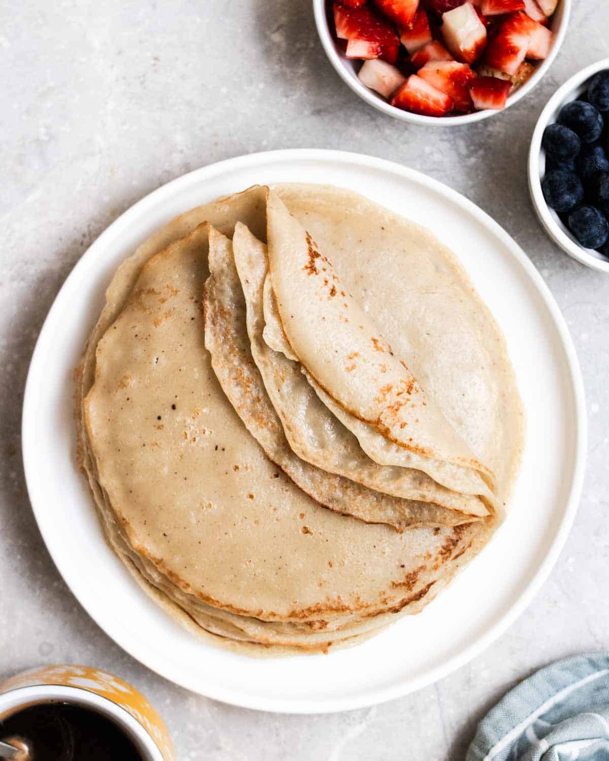 A stack of partially folded crepes on a plate with strawberries, blueberries, and coffee beside.