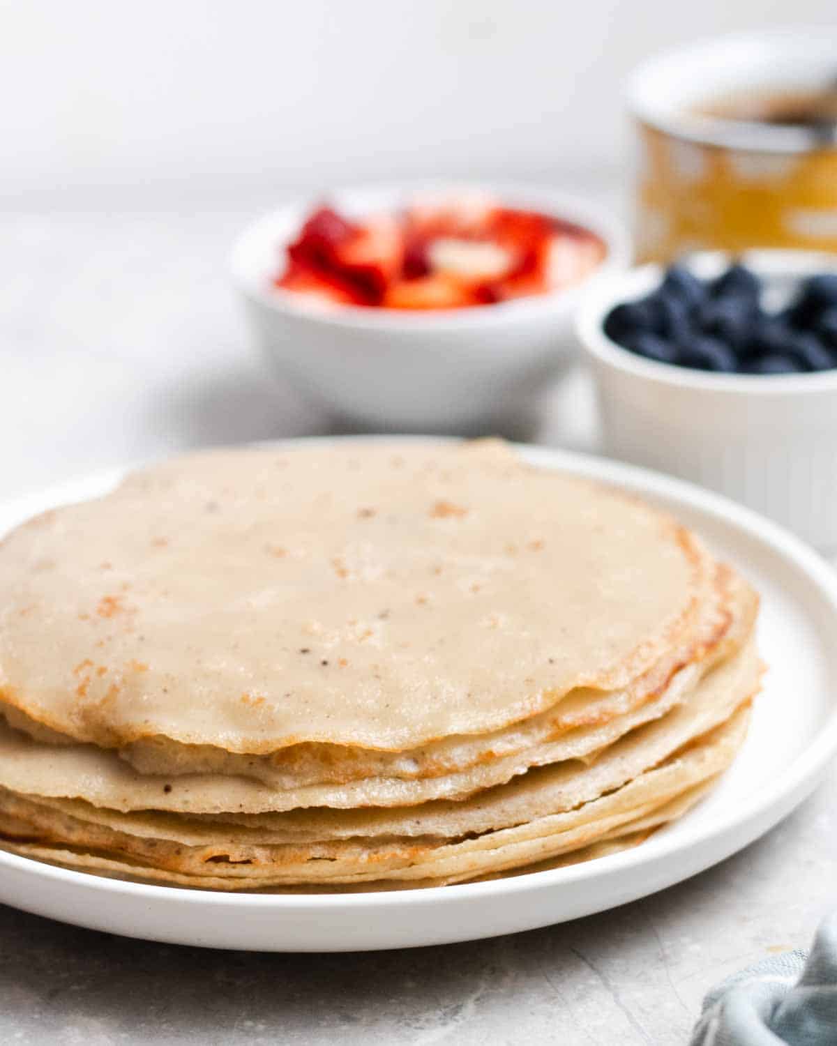 A stack of crepes on a plate with blueberries, strawberries, and coffee in the background.