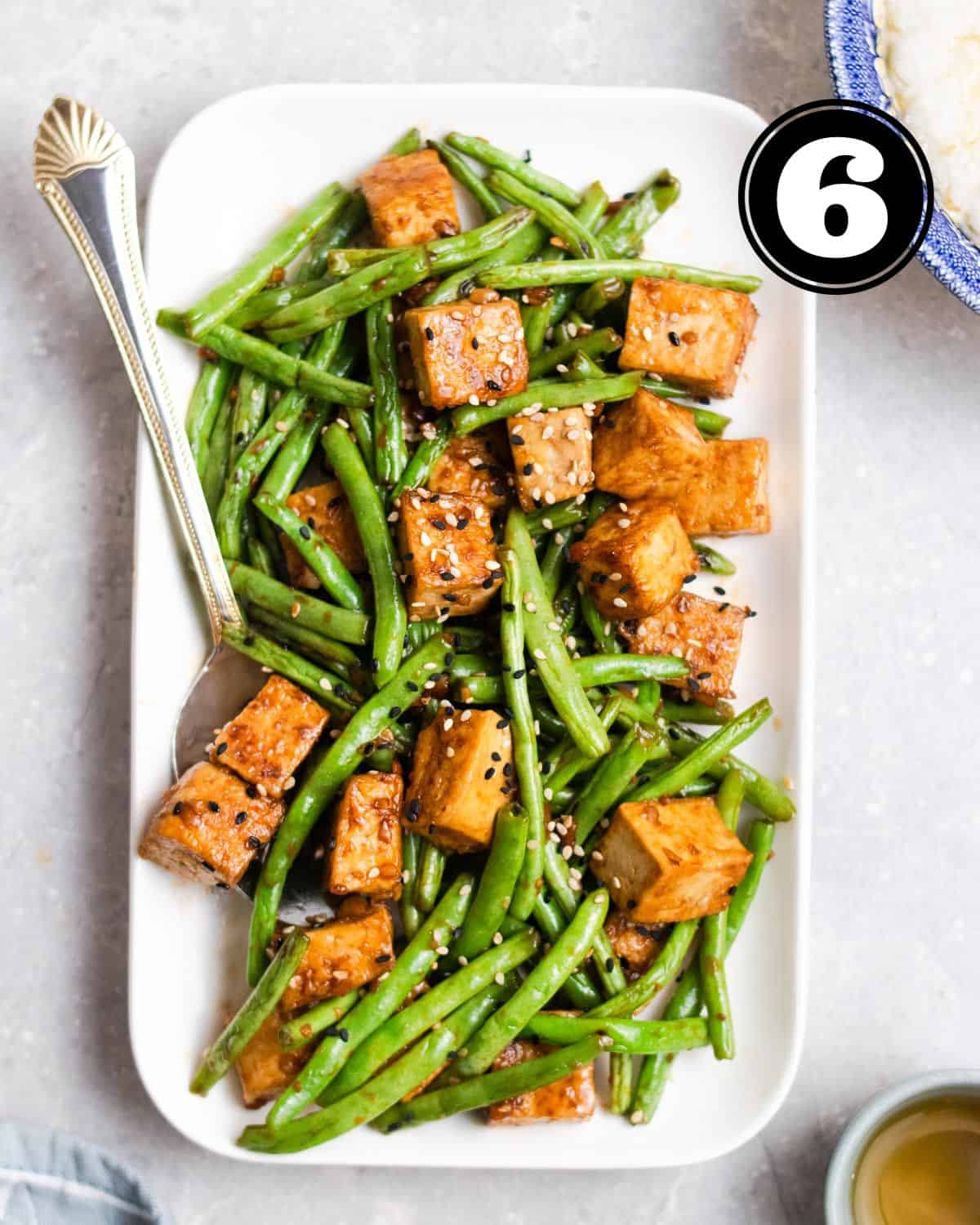 Green beans tofu stir-fry on a rectangular plate served with spoon topped with sesame seeds.