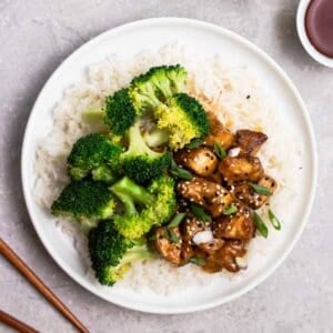 Teriyaki tofu and steamed broccoli served on a plate of rice with chopsticks and cups of tea beside.