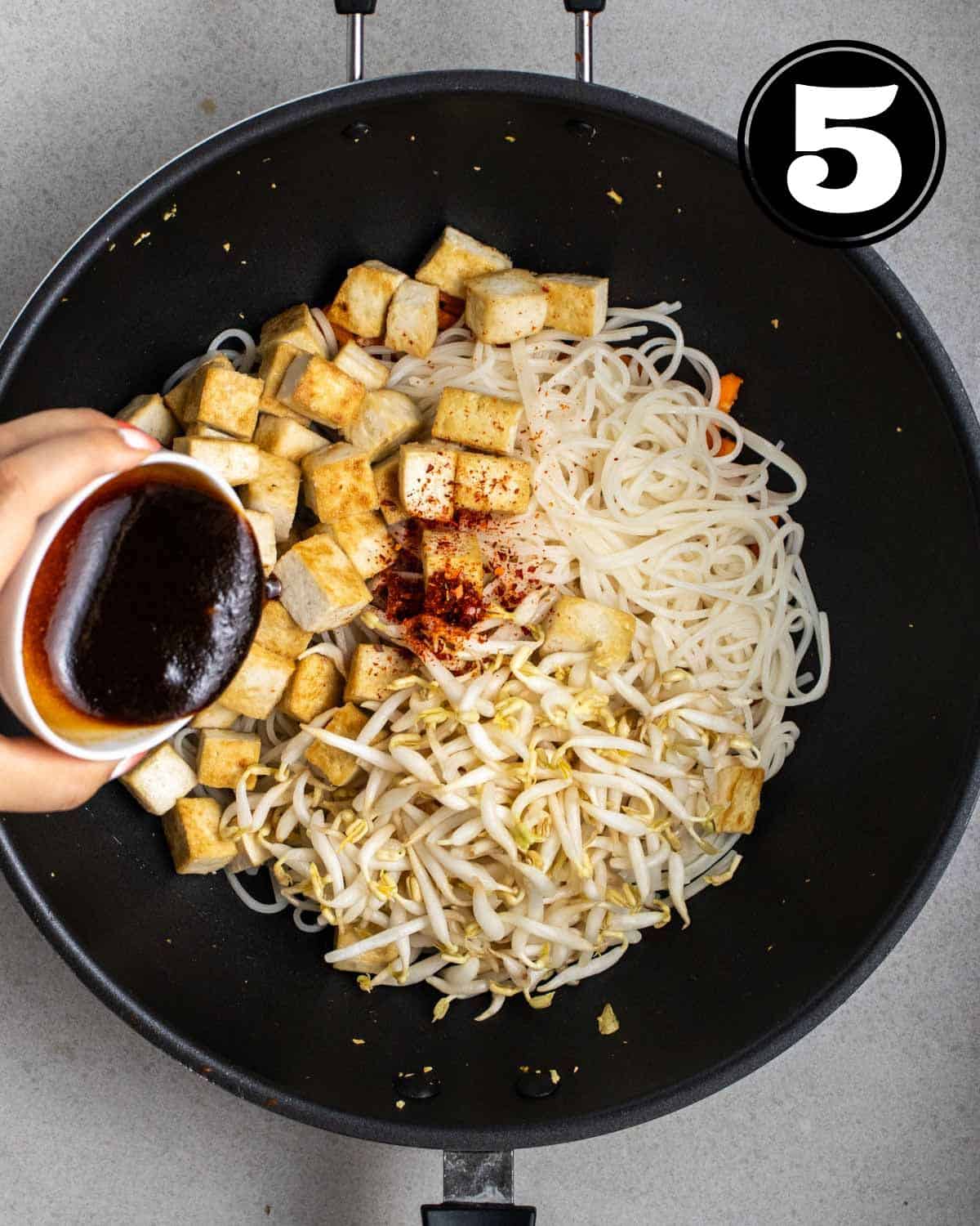 Pouring sauce into a pan of noodles, tofu, and vegetables in a wok.