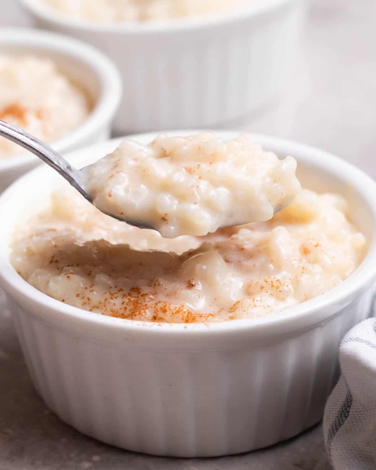 Spooning out some rice pudding from a ramekin with 2 more ramekin of rice pudding in the background.