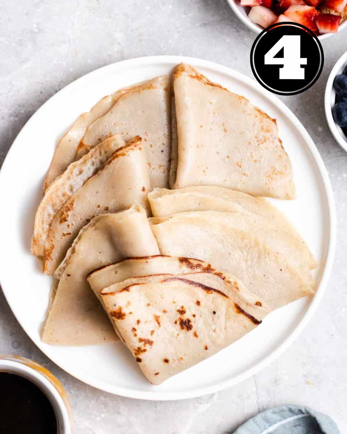 Crepes folded in triangles on a white plate with chopped strawberries and blueberries beside. 
