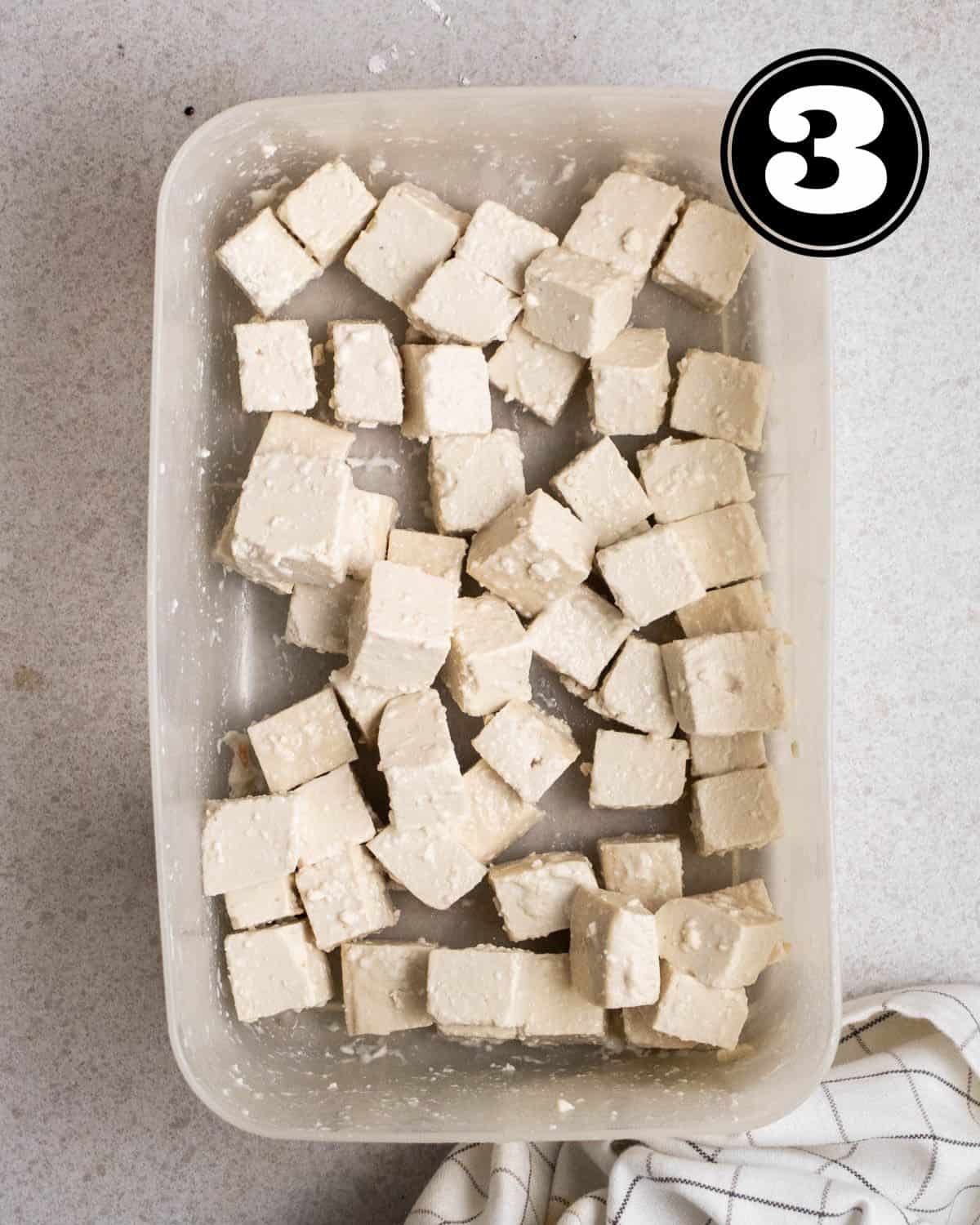 Tofu coated in cornstarch in a plastic container with a white checkered teacloth beside.