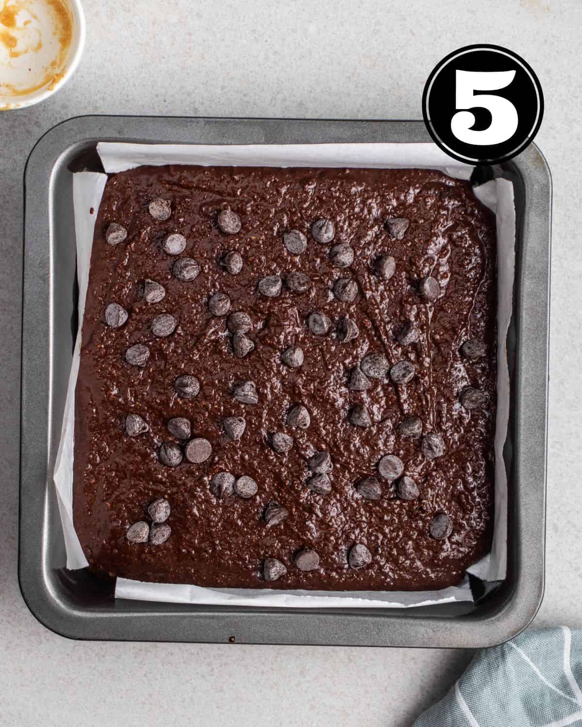 Unbaked brownie in a square pan lined with parchment paper topped with chocolate chips.