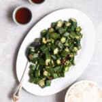 A plate of okra stir-fry served with spoon with 2 cups of tea and a bowl of rice beside.