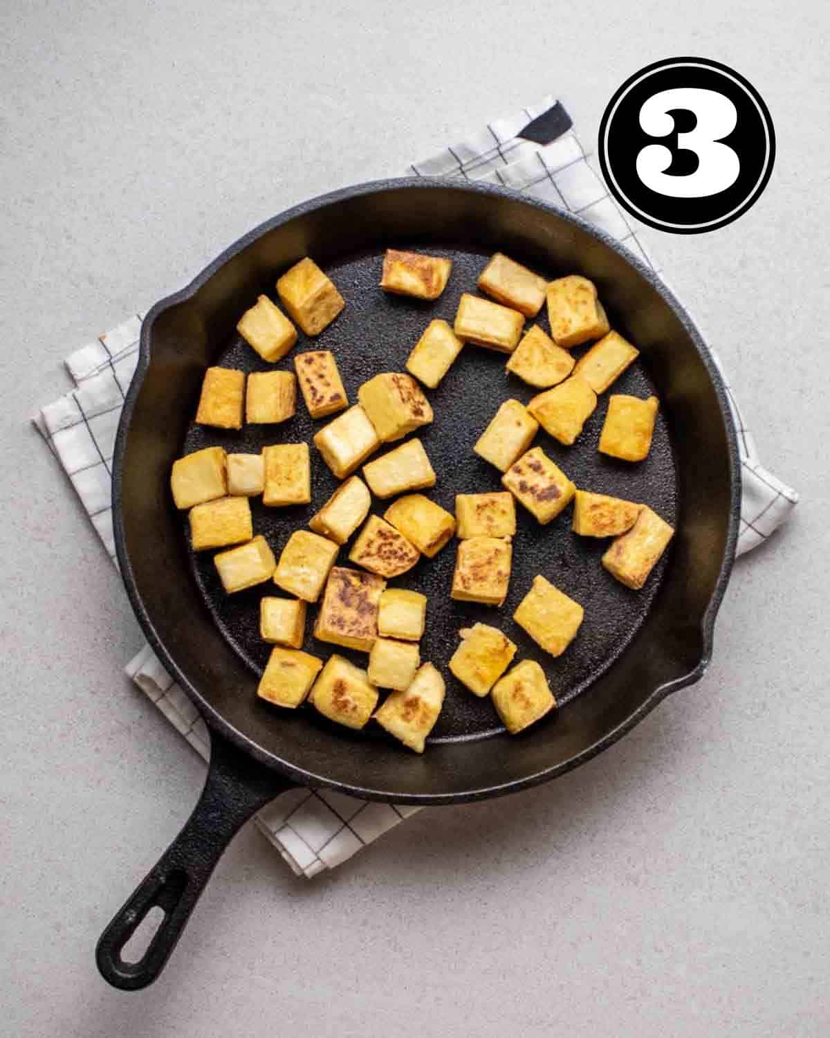 Fried tofu in a cast iron skillet on a white checkered folded teacloth.