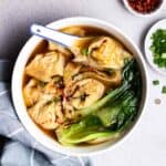 A bowl of wonton soup served with spoon with green onion and chili flakes beside.