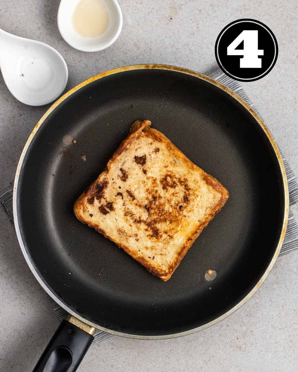A cooked French toast in a non-stick pan on a folded teacloth with 2 empty sauce dishes beside.