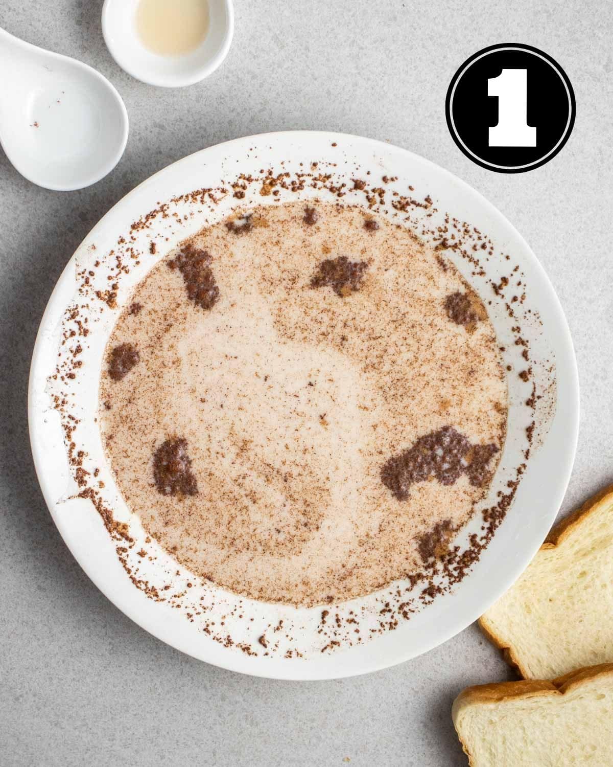 Custard mixture in a white bowl with empty ramekins and 2 slices of bread beside.