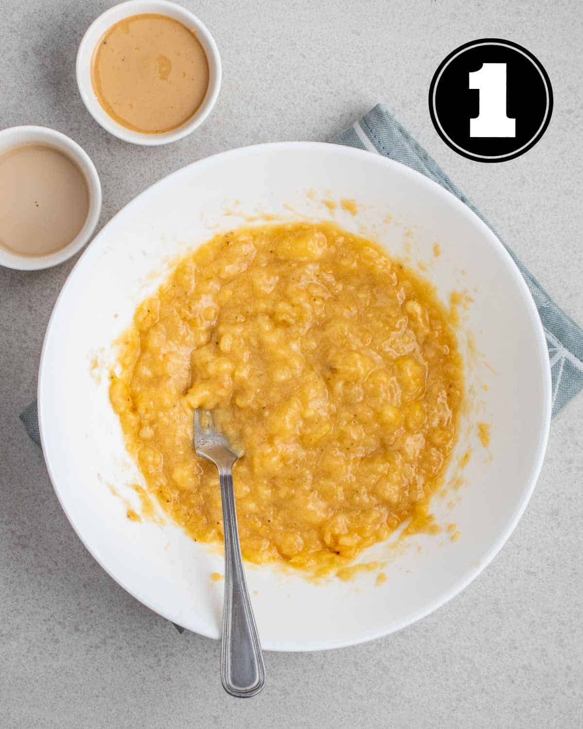 Mashed bananas in a bowl with a fork. There is a ramekin of milk and cashew butter beside.