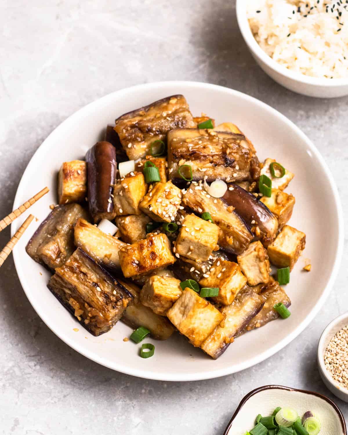 A plate of eggplant tofu stir-fry served with chopsticks with rice, sesame seeds, and green onion beside.
