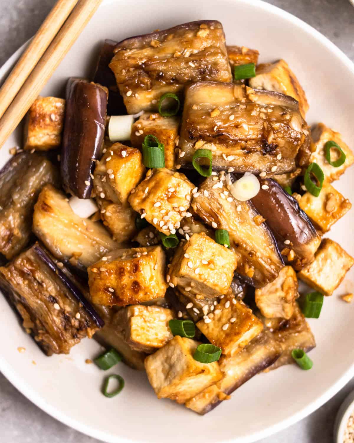 A plate of eggplant tofu stir-fry topped with sesame seeds and green onion served with chopsticks.