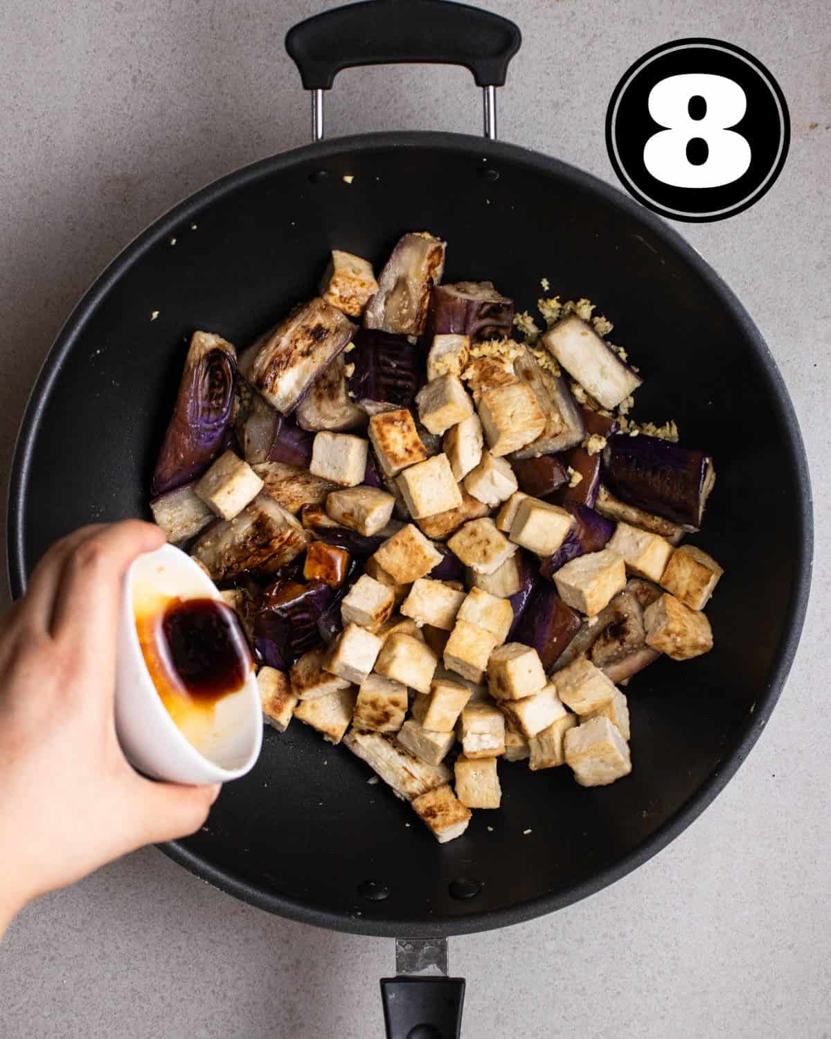 Pouring stir-fry sauce into a wok of cooked eggplant and tofu.