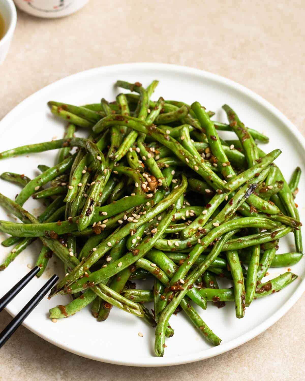 A plate of cooked green beans served with chopsticks topped with sesame seeds.