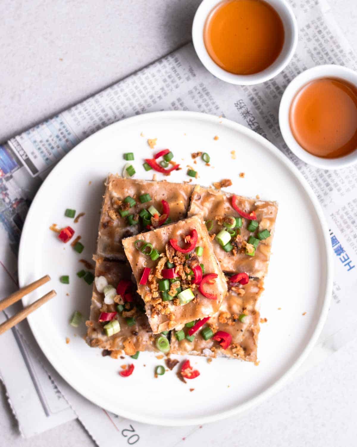 Slices of taro cake arranged on a white plate with a pair of chopsticks and 2 cups of tea beside.
