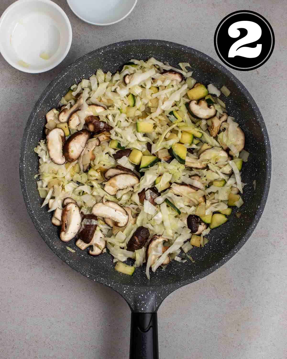 Cooked cabbage, mushrooms, zucchini, and onion in a pan with 2 ramekins beside.