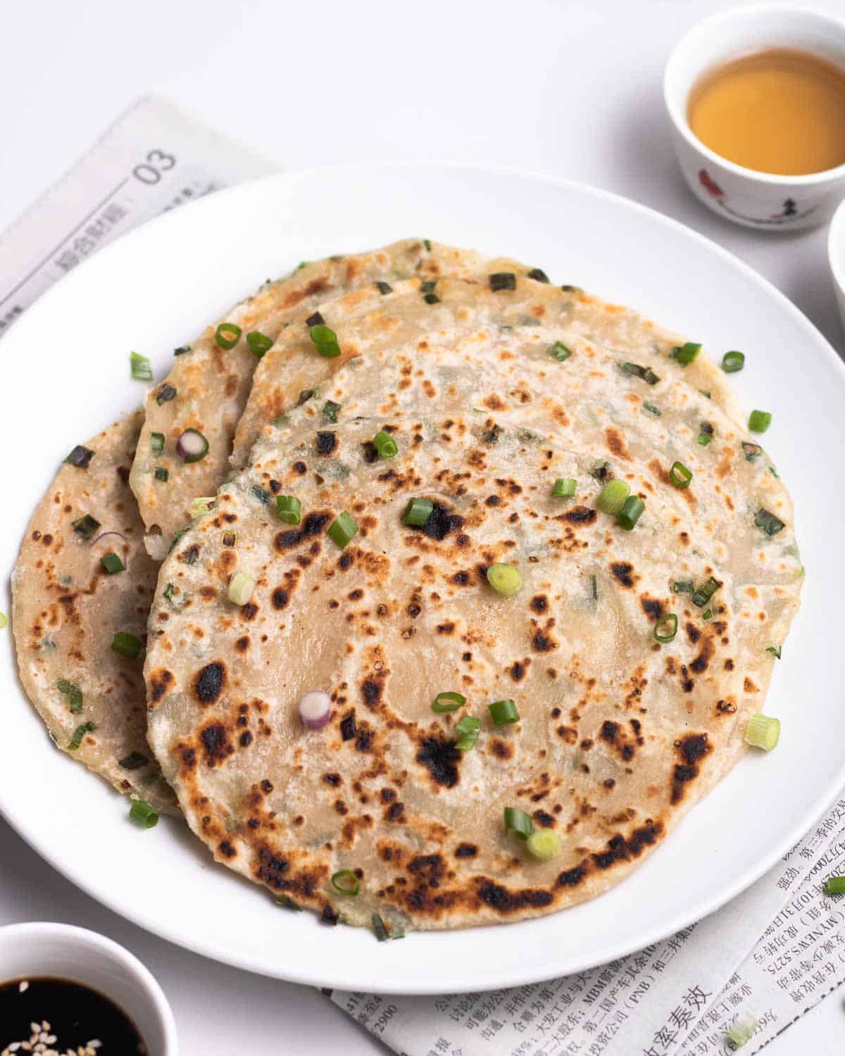 Scallion pancakes on a plate topped with chopped green onion with 2 cups of tea in the background.