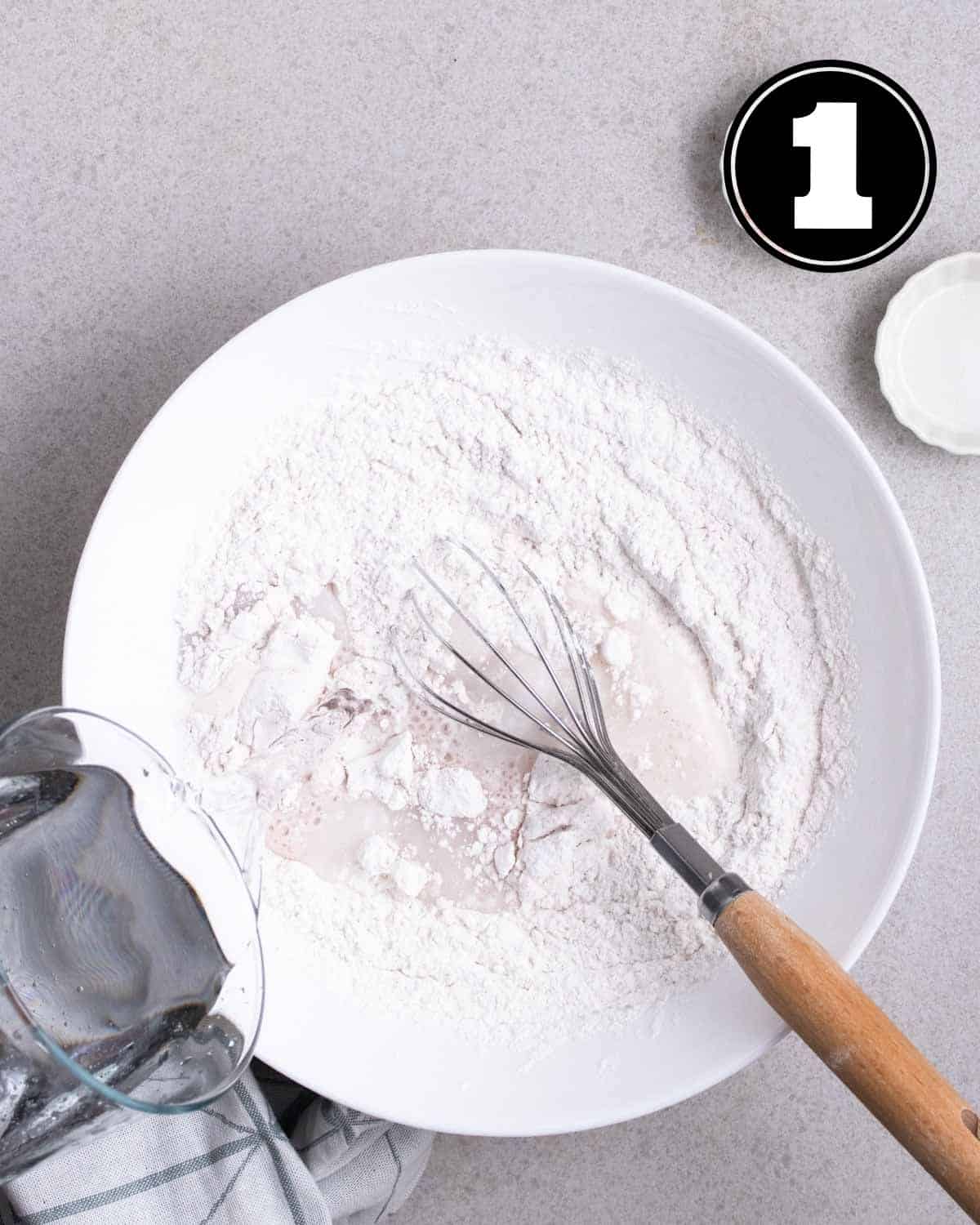 Flour and other dry ingredients in a bowl with a whisk beside.