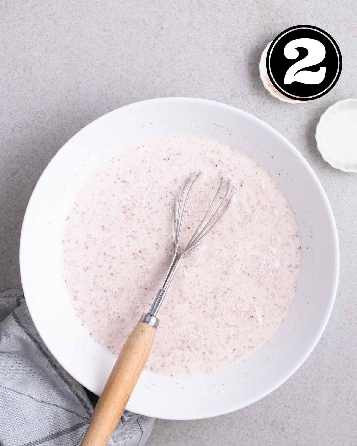 Mixture of flour, seasonings, and water in a bowl with a whisk and 2 small sauce dishes beside.