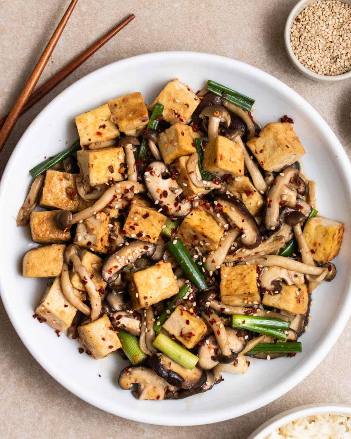 A plate of mushroom tofu stir-fry with a pair of chopsticks, red pepper flakes, and sesame seeds beside.
