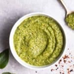 A bowl of pesto with a spoon, basil leaves, and chili flakes in the background.