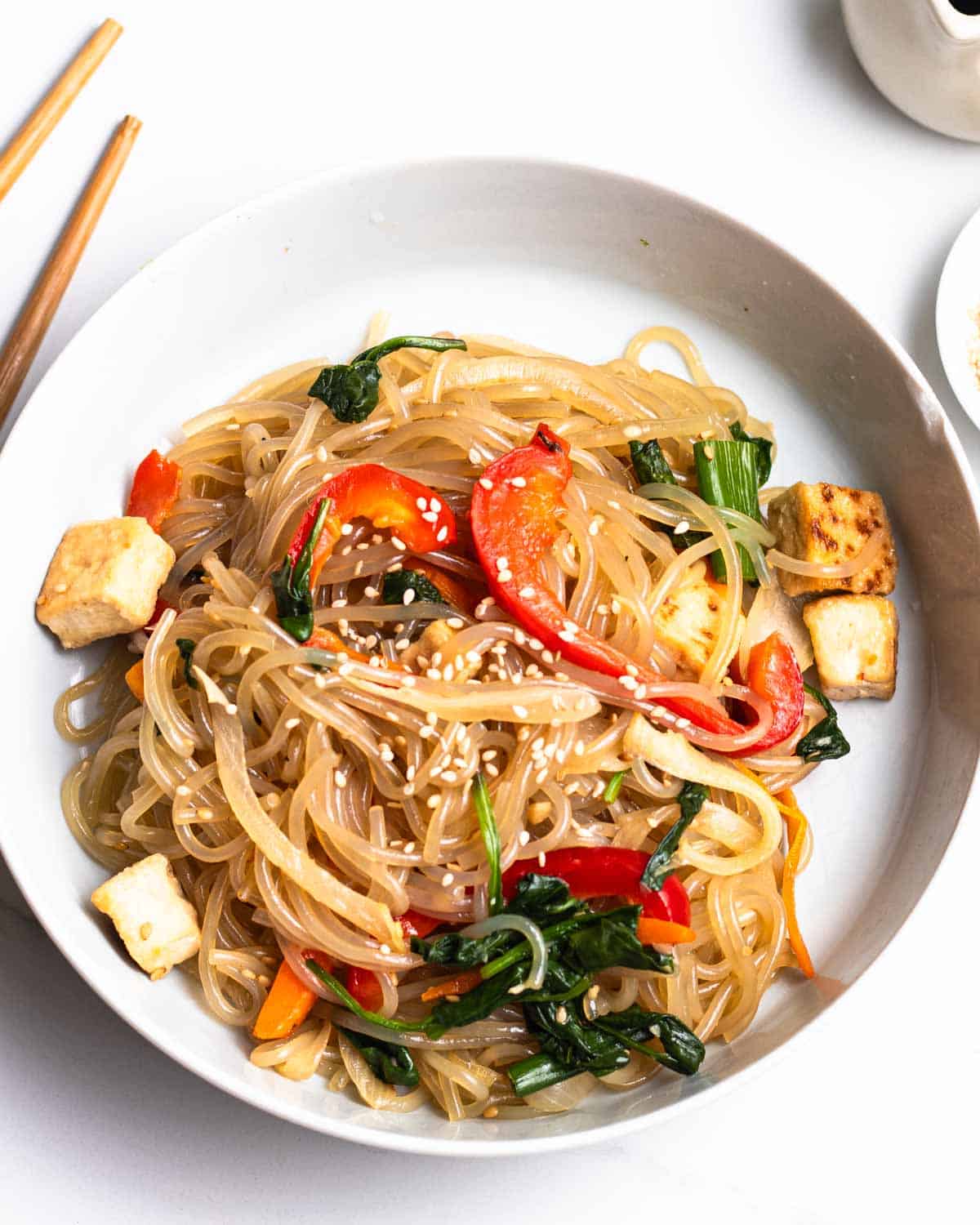 A plate of japchae topped with sesame seeds with a pair of chopsticks in the background.