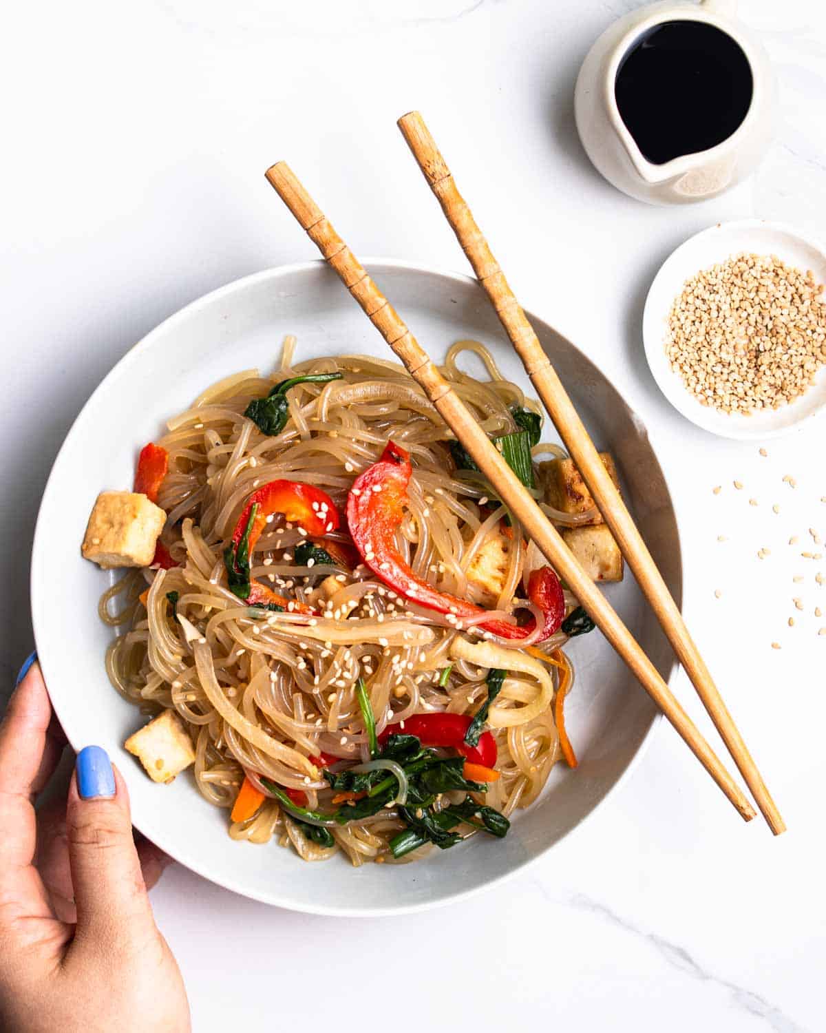 Holding a plate of japchae served with chopsticks with sesame seeds and soy sauce beside.