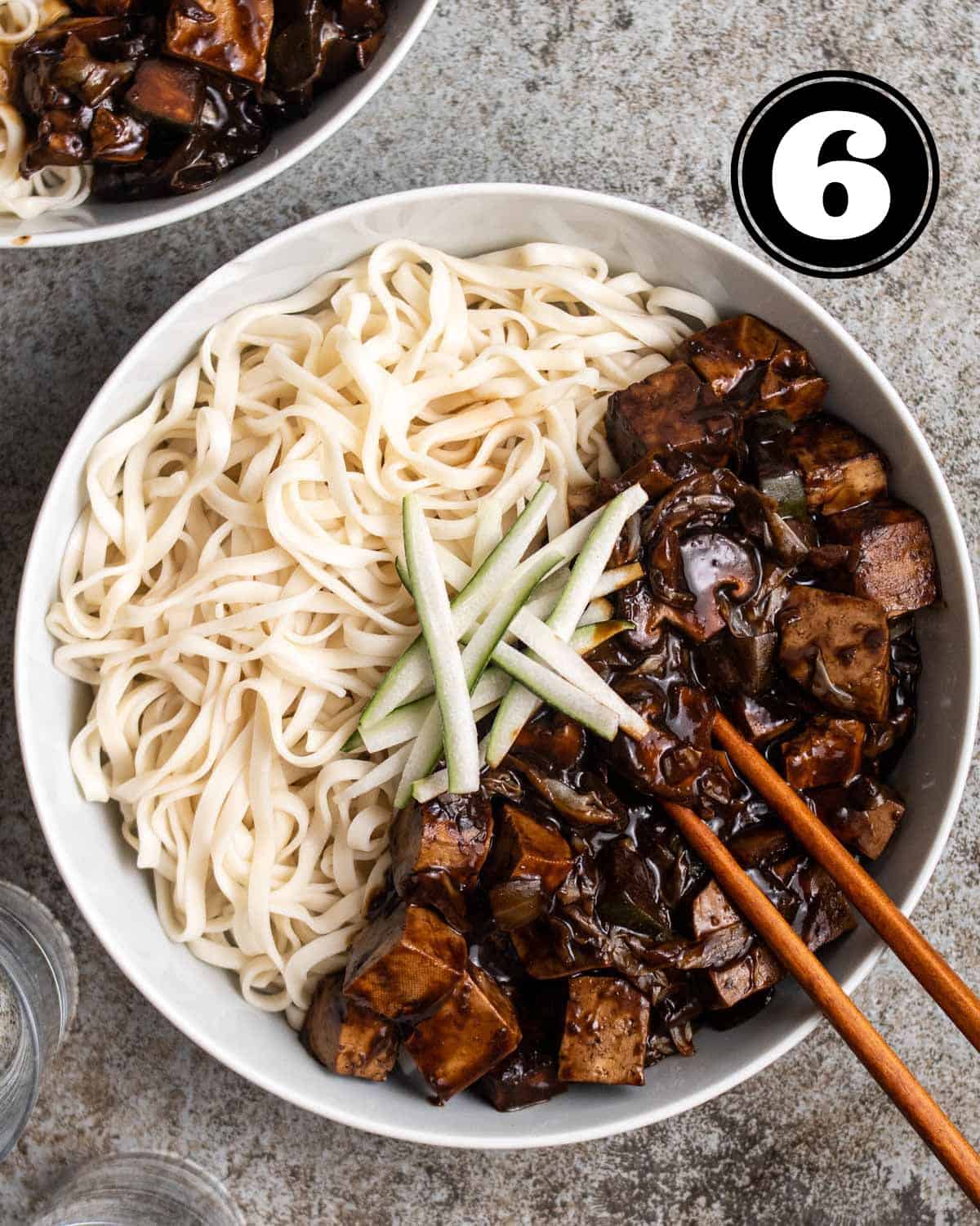 A bowl of jjajangmyeon topped with cucumber slices with a pair of wooden chopsticks beside.