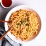 A bowl of peanut butter noodles served with chopsticks with sriracha beside.
