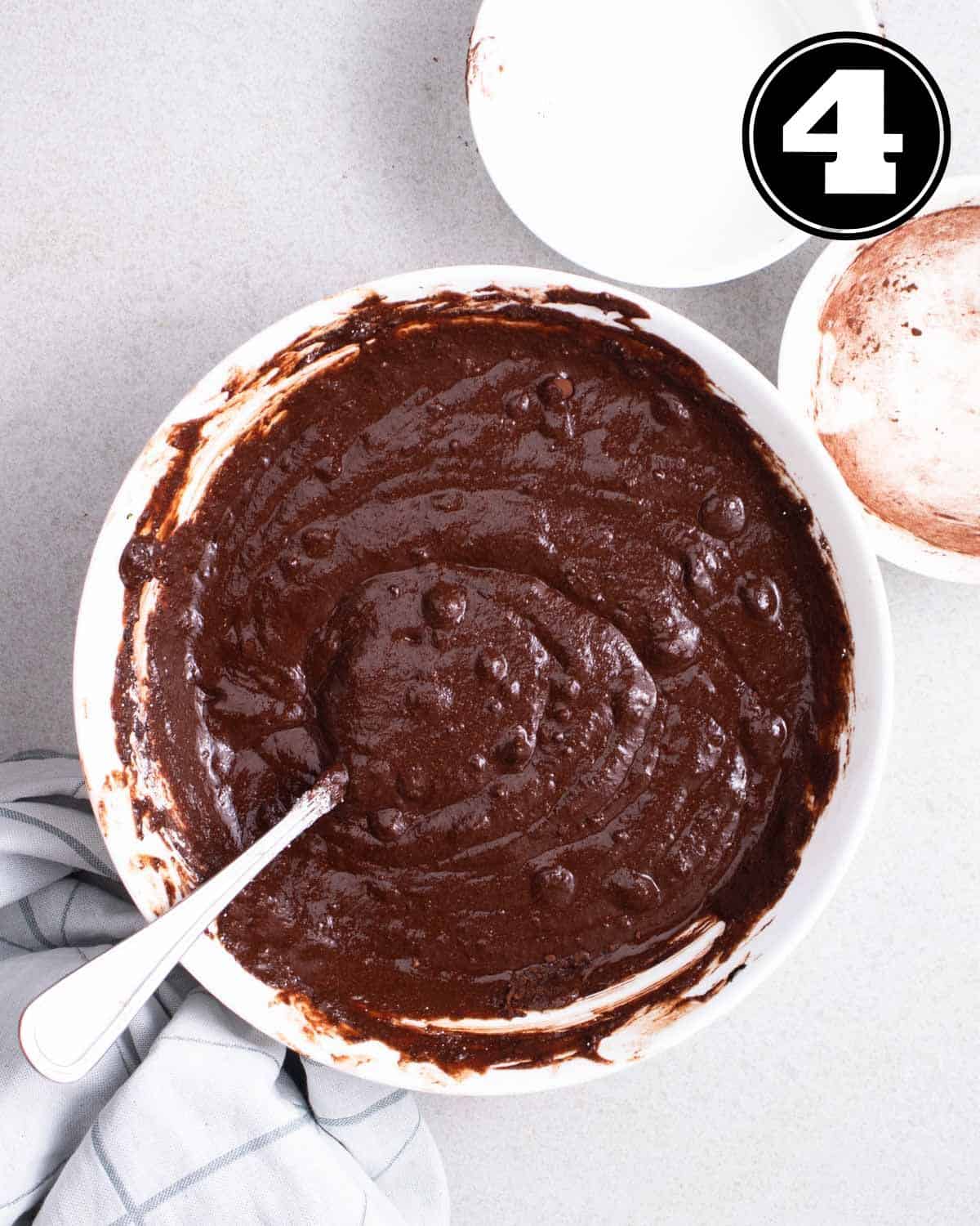 Brownie batter in a bowl with a fork and 2 bowls in a background.