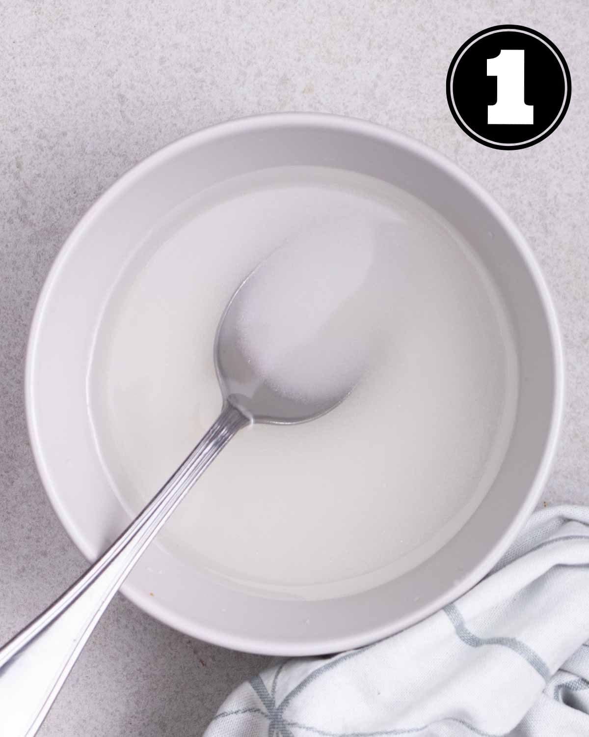 Sugar and water in a bowl with a tablespoon beside.